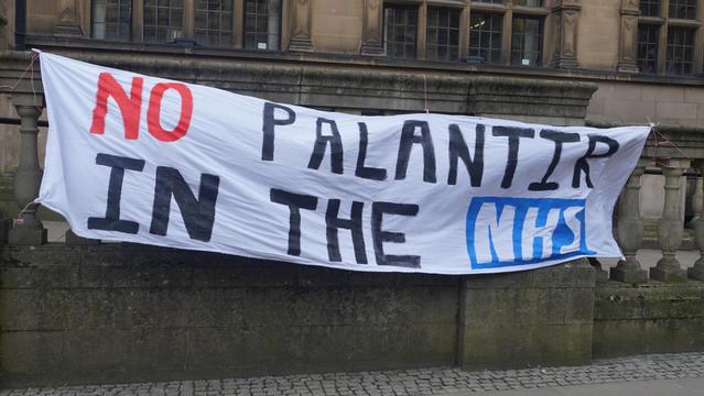 A photo at the No Palantir in the NHS protest outside of Sheffield Townhall a big hand painted banner that says NO PALANTIR IN THE NHS. No is in red, NHS is in the NHS logo.