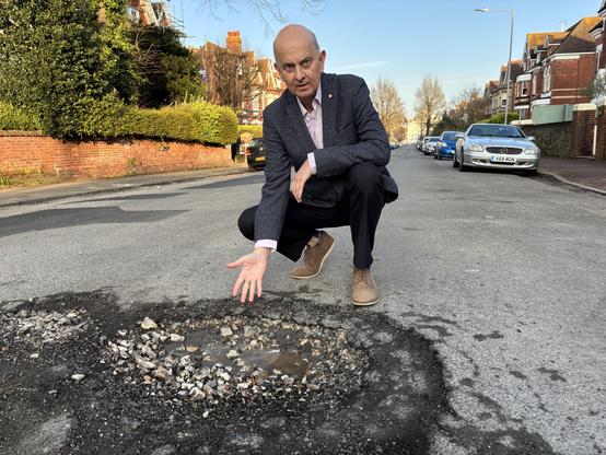 Main in suit jacket crouching behind and pointing at a pot hole. A campaign photo I took (this is Brett Wright, East Sussex County Councillor in Meads, who changed from LibDems to Greens a few weeks before elections were called)