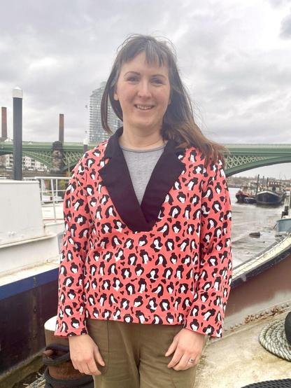 A white woman with straight brown hair is standing on the deck of a barge wearing a sweatshirt made from peachy-pink fabric with a repeating black and white print. It's got a black notched collar and is worn over a grey shirt.