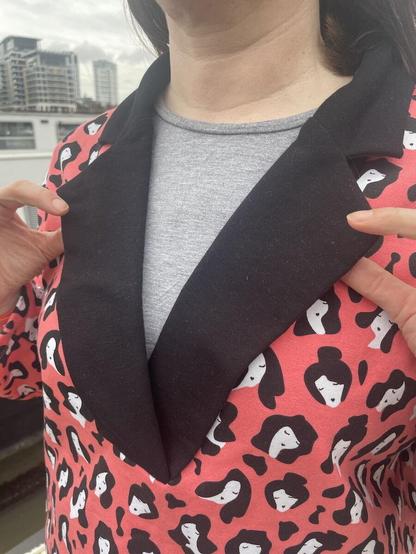 Detail of a sweatshirt made from peachy-pink fabric with a repeating black and white print. It's got a black notched collar which is being held open by white hands, and is worn over a grey shirt.