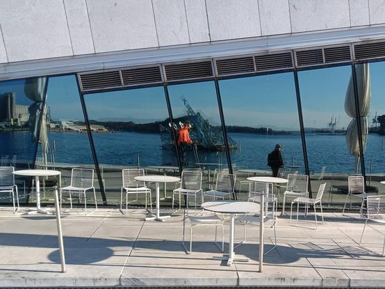 A very blury image of man in a bright orange reflective jacket, riding a tallbike, taking a picture of his reflection in a large glass window. The window area is triangular. In the foreground are white tables and chairs. The Oslo fjord is behind. Directly behind the rider of the tallbike is the sculpture "She Lies".