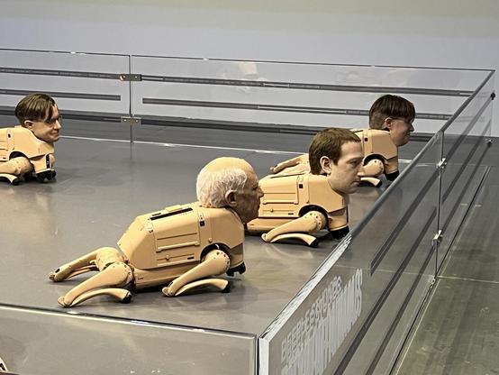 The image features several humanoid robot sculptures with realistic heads and mechanical bodies resembling crab-like designs. The robots are positioned on a flat surface behind a transparent barrier, showcasing a blend of technology and art. One robot resembles Mark Zuckerberg.