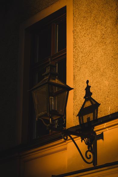A vertical photograph of a partly sunlit yellow plastered wall of an old building. On the wall is a lamp sticking out with an ornate black metal stand and a four sided housing with glass panes around the light source.
The lamp is sitting in sunlight and casts a shadow on the wall behind it.
Behind the lamp is a multi paned rectangular window. The plaster on the wall is mostly rough, but smooth in a frame around the window and on the bottom part of the wall.