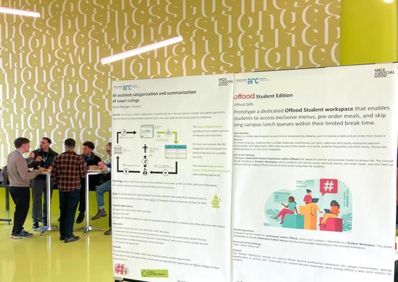 People milling around posters with hackathon challenges. Lots of text and illustrations explaining projects for charities.