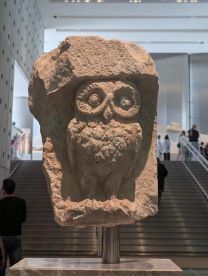 A carved stone owl in a dedicated to the goddess Athena in a display case at the Acropolis Museum framed by stairs leading up