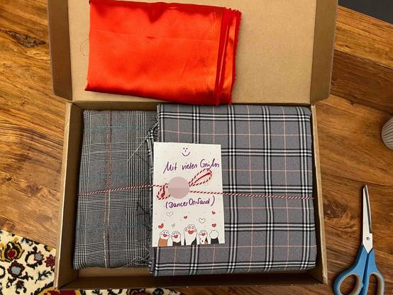 A box contains three pieces of fabric: one red satin and two plaid. There is also a card with a cheerful message attached. A pair of scissors sits nearby on a wooden surface.