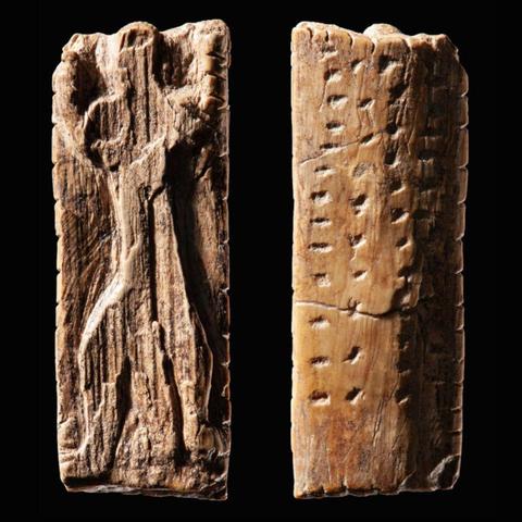 The image displays the Adorant, a prehistoric figurine carved from mammoth ivory. It is a small ivory plaque depicting a human-like figure with arms raised in a pose of adoration or worship. The reverse side features orderly sequences of notches and dots, which researchers suggest may represent an early precursor to written language or a structured communication system.