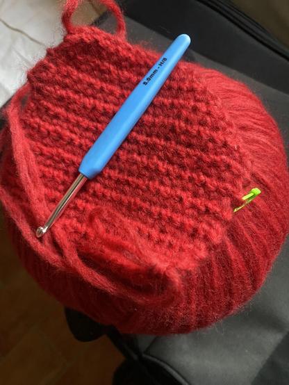 A blue crochet hook resting on a swatch of single crochet which is resting on a large ball of soft red yarn.