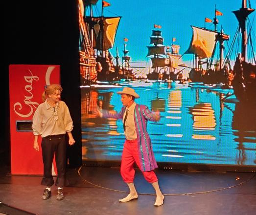 Photo from the Monkey Island stage play in Halle an der Saale, Germany. Stan, flailing around with his arms, as he does, is trying to sell Guybrush the "Sea Monkey". In the background, there's a big screen showing a backdrop with a few ships. To the side, there's the iconic Coca-Cola styled Grog vending machine.