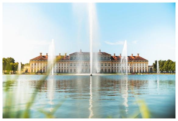 Blick über ein Gewässer mit Springbrunnen; dahinter eine breite Schlossanlage mit gelb/weißen Mauern und daneben Bäumen.