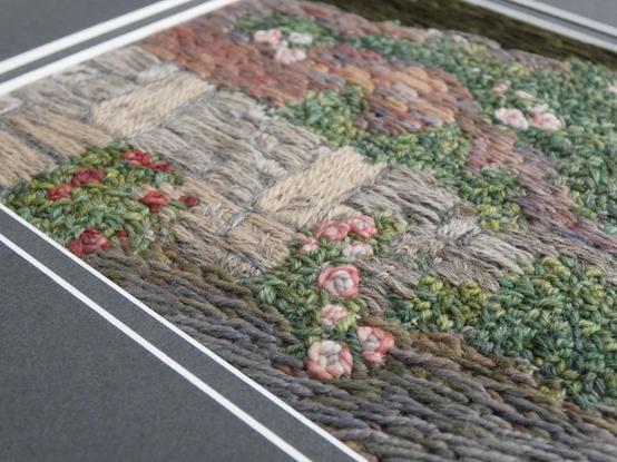 Part of a stitched artwork depicting roses growing up a stone wall. Viewed on an angle to show texture