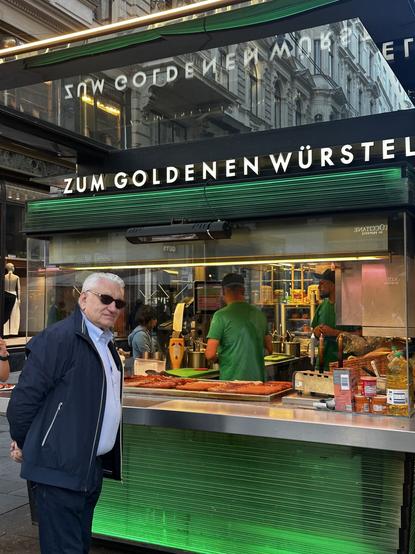 Austria’s finest cuisine a sausage stand. Green backlighting and a suit wearing upper class customer beside it.
