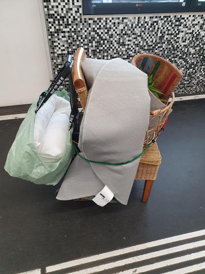 Eine Sackrodel über und über bepackt mit einem Sessel, einem Teppich, einen Fußvorleger, einem Korb, einer Pflanze und einem mit einem Polster gefüllten Sack. Die Sackrodel ist kaum zu sehen. Das ganze Ding steht auf einem Bahnsteig und wartet auf die Straßenbahn.