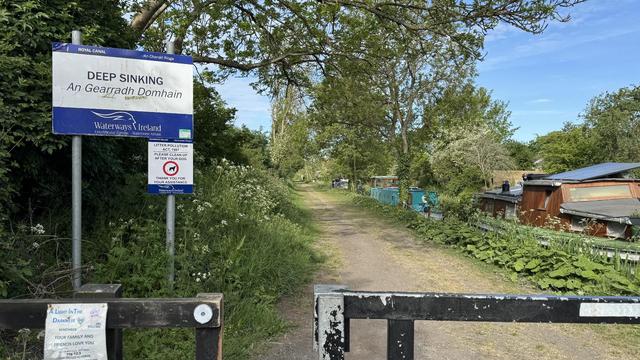 Photo, landscape, exterior: A canal towpath, trees and undergrowth on the left, canal with boats on the right, a gate in the foreground, a sign indicating ‘Deep sinking’. It’s a bright day.