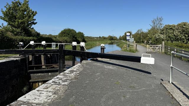 Photo, landscape, exterior: A canal lock to the left of the towpath/greenway. Trees in the distance on the left. It’s a bright day.