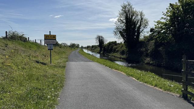 Photo, landscape, exterior: A canal towpath/greenway. Canal to the right, trees beyond. A sign indicates that this is a ‘Shared space’. It’s a bright day.