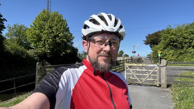 Photo, landscape, exterior: A selfie of a middle-aged bearded man in red, white, and black cycling gear and helmet. He is a gravel cycling hero, you can tell. Beyond is a fence. That’s right, it’s a bright day.