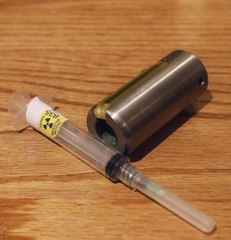 A syringe with radiation warning sticker next to a thick metal tube it goes inside