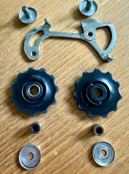 part of derailleur pulley wheels pulled off, both look similar but the one on the right has slightly thinner teeth (and says "NARROW"). The one on left also has extra ring of metal in the center instead of plastic, making it look like it can handle more load. Below them are little still cylinders that go into center of wheels. Also show metal discs to keep the bearing areas covered on both sides and part of derailleur they screw into.