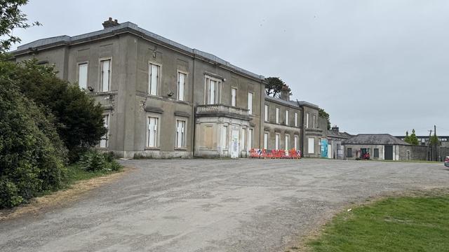 Photo, landscape, exterior: A large, dilapidated country house, windows and doors boarded up. Some bushes to the left, and some temporary builders barriers in front, outhouses to the right (in better condition).