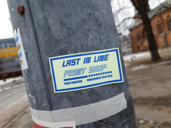 A rectangular light yellow sticker on a metal pole with a blue border. Blue text reads "LAST IN LINE PRINT SHOP" above the name "TRULS RINGSTAD" and address "BIRKELUNDEN, 0553 OSLO".