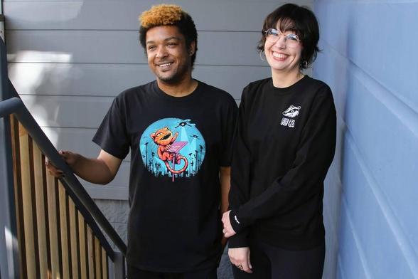 Two people stand on a staircase, one wearing EFF's new Claw Back tee featuring an orange cat climbing an ALPR camera to swat at surveillance drones in a blue night sky. The second wearing EFF's new Privacy Badger Crewneck sweatshirt. Black sweatshirt with a white cartoon badger on the chest, above characters that say "privacy".