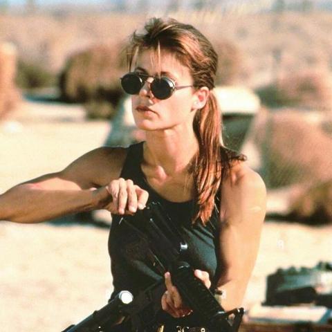 Linda Hamilton as buffed-up Sarah Connor in T2 wearing a pair of sunglasses,  a ponytail, and a dark tank top that showcases her toned arms. Overall looking very badass and taking no nonsense.