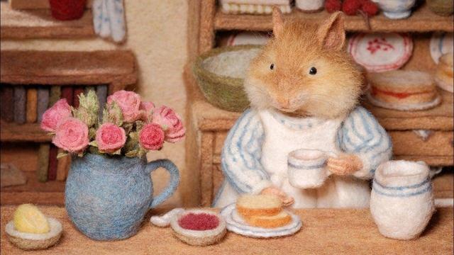 A felted wool hamster character wearing a white apron sits at a kitchen table with a pitcher of pink roses, a cup of tea, and small dishes of bread and jam.