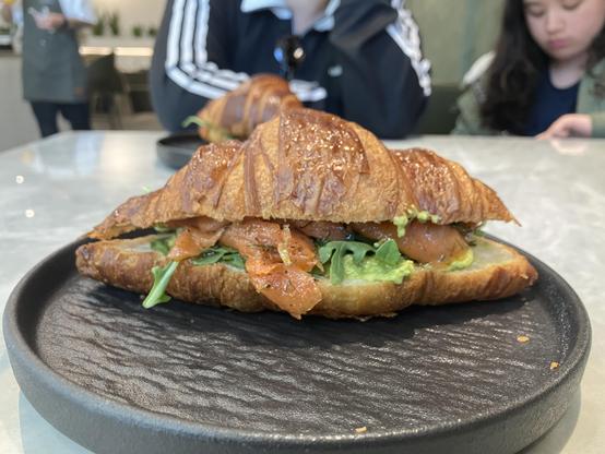 A flaky croissant sandwich sits on a matte black ceramic plate on a café table. The croissant is deeply golden and glossy, filled with slices of smoked salmon, green avocado spread, and fresh arugula peeking out from the sides. The foreground is sharply focused on the layered pastry and filling, while the background is softly blurred, showing two children seated at the table and another croissant further back. The café interior has muted lighting and a modern, casual atmosphere.
