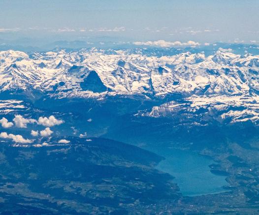 A aerial view from the north looking down the Thunersee in Switzerland towards the snowy Alps, with the black triangle of Eiger Nordwand standing out against the sunlit mountains around.