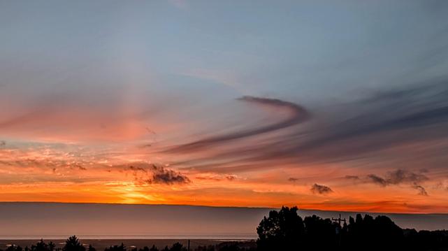 The sun is disappearing behind a fog bank over the ocean. The light from the fading sun looks like a beacon shining straight up. The clouds are curvy.