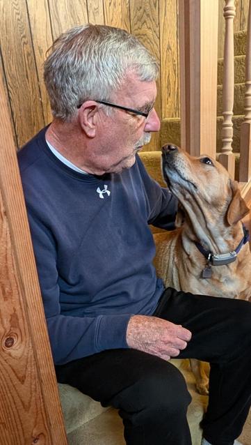 An older man in glasses who needs a shave. He is playing with his Golden Labrador Retriever while sitting at the bottom of a staircase.