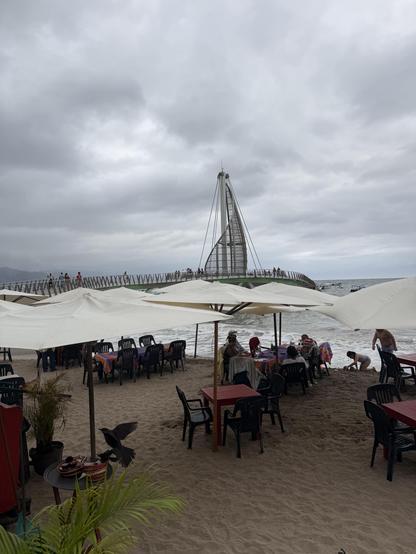 The conical sail pier in Puerto Vallarta’s Zona Romantica as seen across the umbrellas of a restaurant along the boardwalk. The sky is cloudy but bright.