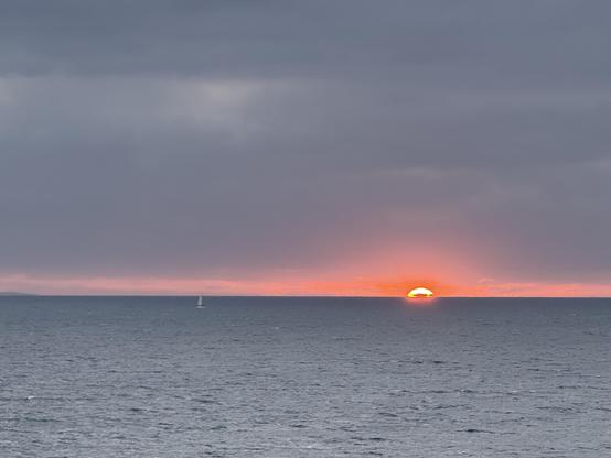 A moody gray sea and matching sky sandwich a fiery sunset as the sun dips its last third below the horizon. A distant sailboat balances the composition