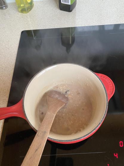 A red pot on a stovetop contains a simmering mixture of porridge & muesli, with steam rising. A wooden spoon is resting inside the pot. In the background, there are bottles of oil and a spice container.