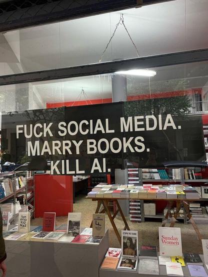 A storefront window of a bookstore displaying a large black sign with white text that reads, "FUCK SOCIAL MEDIA. MARRY BOOKS. KILL AI." Inside the store, wooden tables and shelves are filled with various books. Reflections of the street and buildings are visible on the window glass.