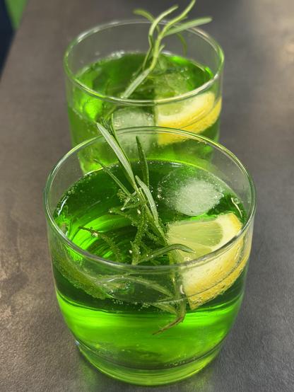 Leuchtend grüne Drinks mit Eiswürfeln, Zitrone und Rosmarin in Gläsern auf einer Arbeitsfläche.
