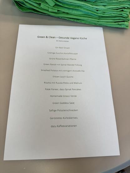 Eine Menükarte für „Green & Clean – Gesunde Vegane Küche“ liegt auf einem Tisch, daneben ein Stapel grüner Schürzen oder Tücher.