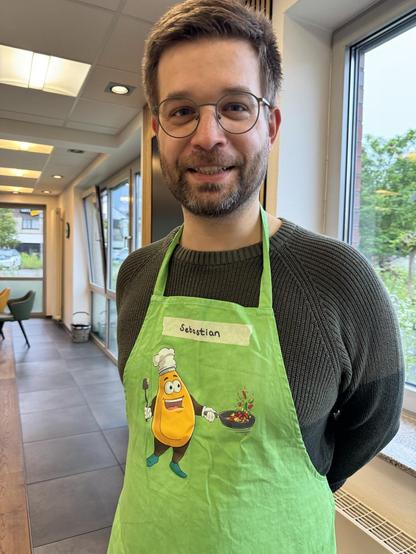 Eine lächelnde Person mit Brille trägt eine grüne Kochschürze mit Namensschild „Sebastian“ und einem Cartoon-Gemüse als Kochmotiv. Im Hintergrund ist ein heller Raum mit großen Fenstern zu sehen.