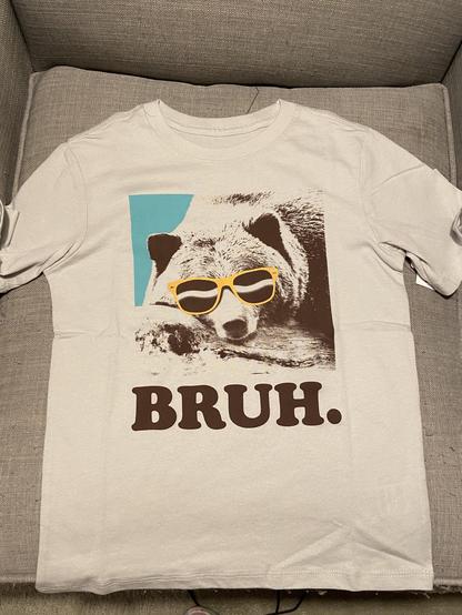 A white short-sleeved t-shirt is laid flat on a gray textured couch. The shirt features a graphic print of a bear's head wearing bright yellow sunglasses. A light blue rectangular shape is visible behind the bear graphic. Below the image, the text "BRUH." is printed in large, bold, dark brown capital letters.