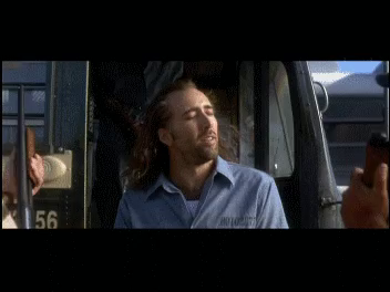 Nicolas Cage in CON AIR, standing in the open and smiling broadly as the breeze blows his long hair.