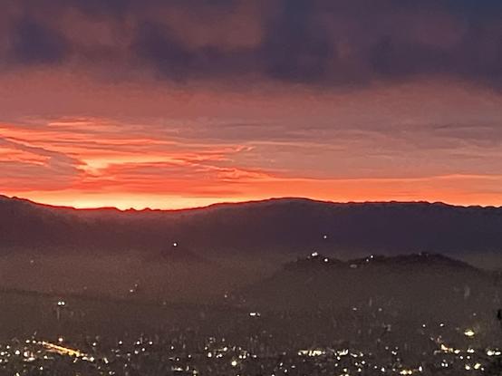 The hilltops of the coastal mountain ranges that surround Santiago on the east, and the city below. It’s sunset and the sky is purple with bright orange glows, and the city lights are on. On a hill you can see the smog clouds reaching just below the top.