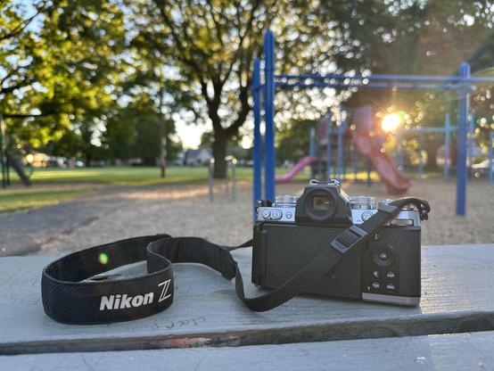 An iPhone picture of a Nikon Z fc camera sitting on a park bench, sunset in the background and light filtering through the trees and the children's playground.