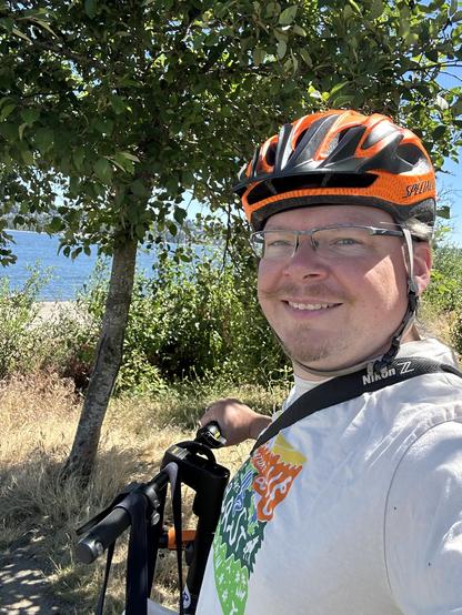 A selfie of me wearing an orange bicycle helmet and standing with my e-scooter. Tall grasses, trees, and the river are directly behind me.