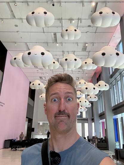 A man attempts to imitate a cartoonish face adorning a series of suspended cloud art characters behind him at a museum.