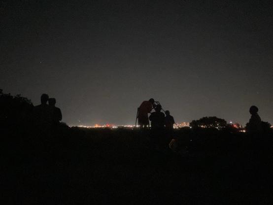 silhouetted figures looking through a homemade telescope against a clear night sky above the boston skyline