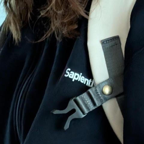 Nahaufnahme einer Person in einer schwarzen Jacke mit einer teilweise sichtbaren weißen Textstickerei "Sapient". Ein beiger Gurt mit einer dunkelgrauen Kunststoffschnalle und einer goldenen Metallniete verläuft über die Schulter. Oben sind braune Haare zu sehen.