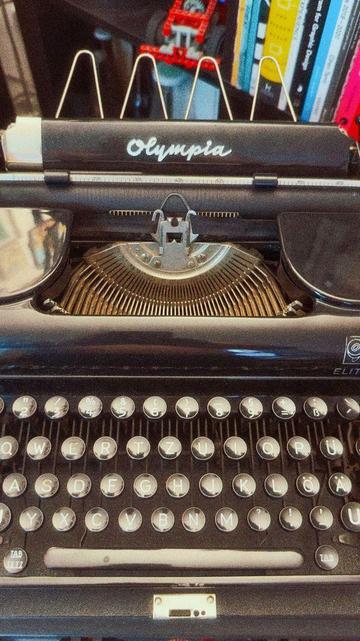 Close-up of an Olympia Elite typewriter keyboard and carriage, with a stack of books and a red construction toy in the blurred background.