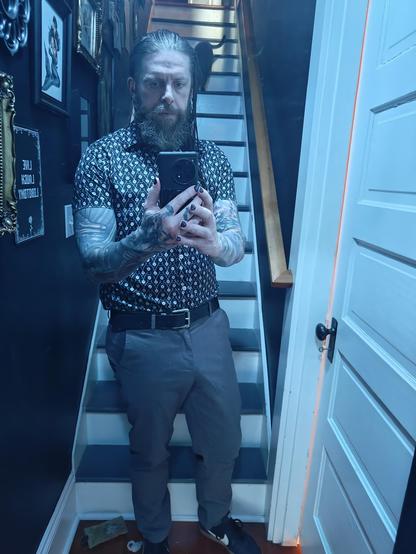A mirror selfie. I am wearing grey slacks, with a short sleeve black button up shirt with a white pattern, tucked in with a belt.