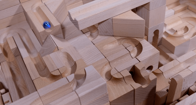 An intricate wooden marble run made of interlocking blocks with curved and straight carved tracks, featuring a single glossy blue marble rolling down a channel on the upper left.